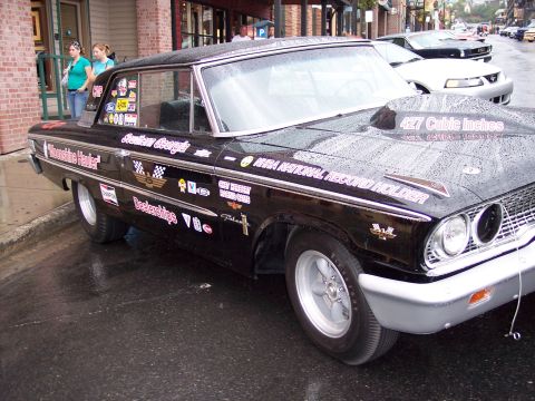 1972 Ford Ranchero - A black 1964 Ford Falcon drag car is parked on a wet street, adorned with numerous racing decals.