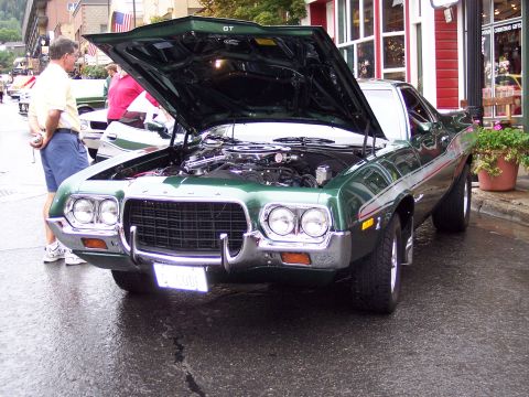 1972 Ford Ranchero - A man looks at the engine of a green 1972 Ford Ranchero parked on a wet street in front of shops.