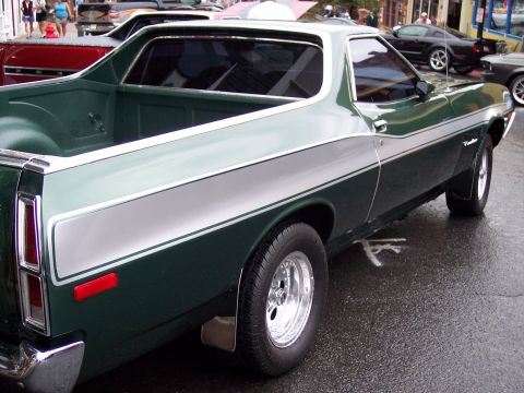 1972 Ford Ranchero - A green pickup truck with a silver stripe is parked on a wet street, with other vehicles and people in the background.
