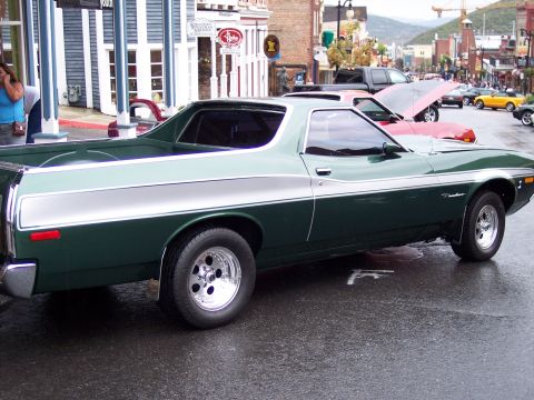 1972 Ford Ranchero - A green 1972 Ford Ranchero is parked on a wet street in a town with shops and mountains in the background.