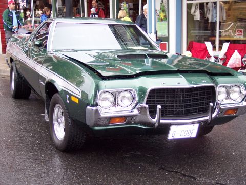 1972 Ford Ranchero - A green 1972 Ford Ranchero is parked on a wet street in front of storefronts.