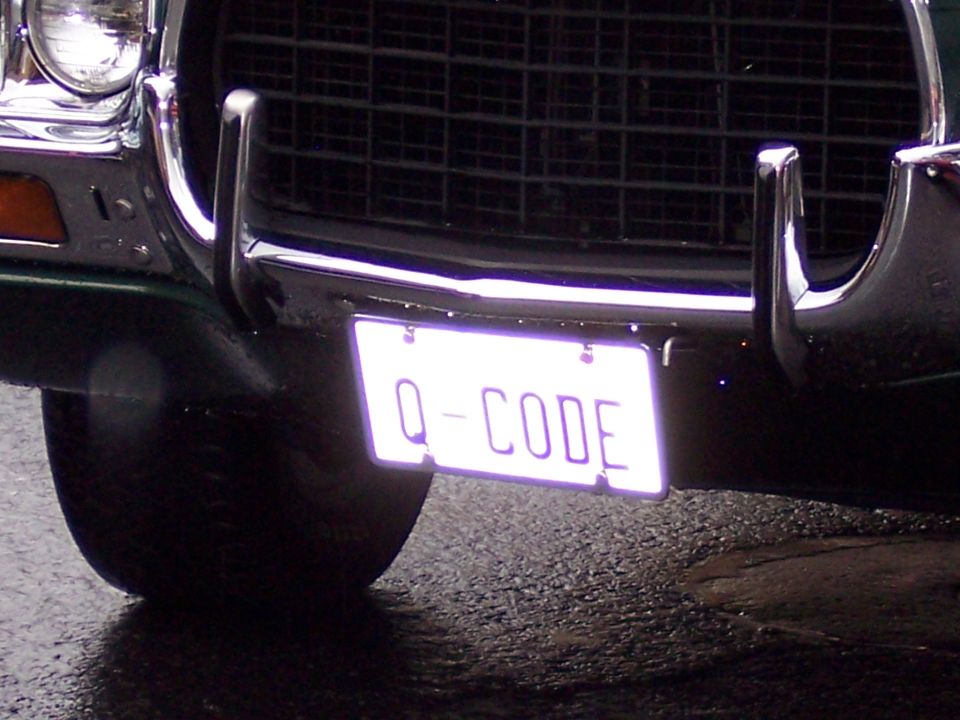 Front chrome bumper of dark classic car with white 'Q-CODE' license plate on wet pavement.