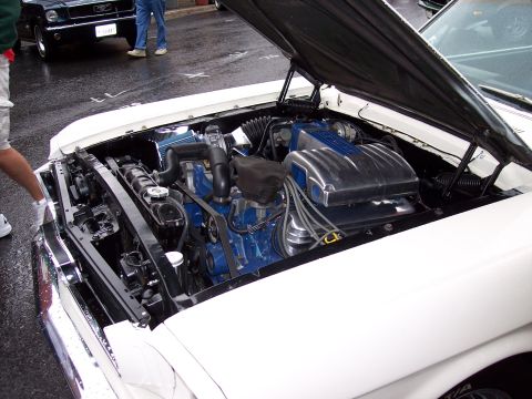 1972 Ford Ranchero - The engine bay of a white vehicle is open, revealing a blue engine block and chrome intake manifold. A black Ford Mustang is visible in the…