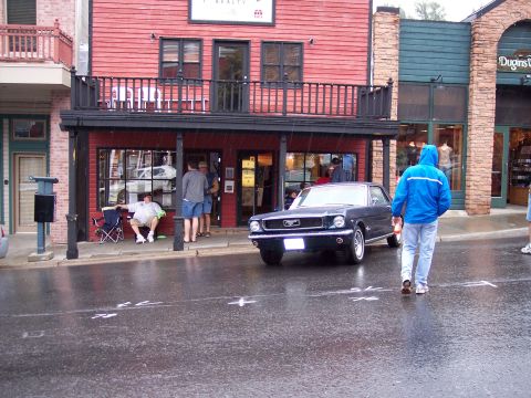 1972 Ford Ranchero - A dark blue 1965 Ford Mustang coupe is parked on a wet street in front of a red building.