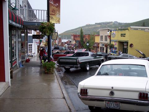 1972 Ford Ranchero - Several cars are parked on a wet street in a town with shops and hills in the background.
