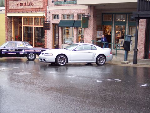 1972 Ford Ranchero - A silver coupe is parked on a wet street next to a black car in front of shops.