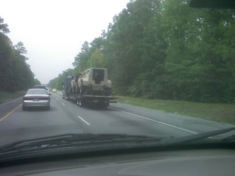 2001 Ford F-150 - A flatbed truck carries a military vehicle down a highway surrounded by trees.