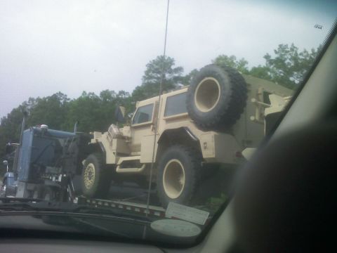 2001 Ford F-150 - A tan military vehicle is loaded onto a flatbed truck, with a blue semi-truck visible to its left.