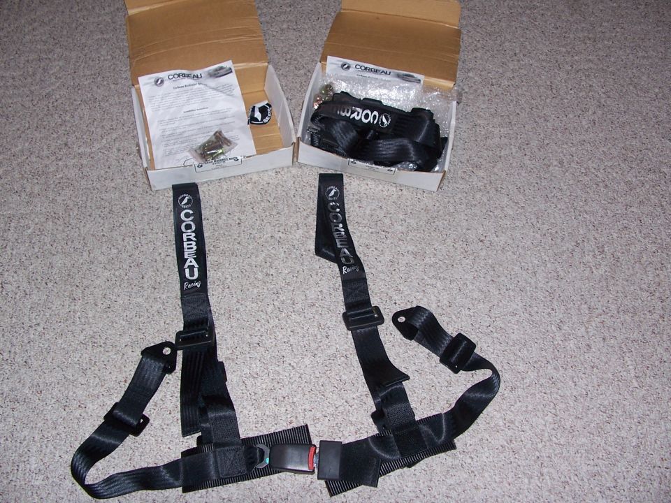 Black Corbeau Racing seat harness belts unboxed on carpet with instructions and hardware