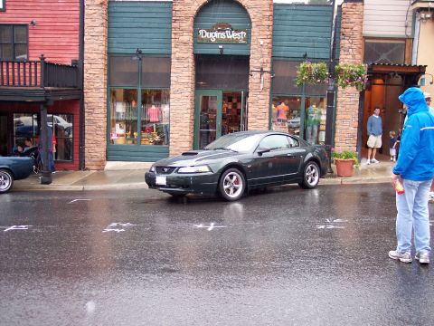 1972 Ford Ranchero - A dark green Ford Mustang is parked on a wet street in front of a store, with another vehicle partially visible to the left.