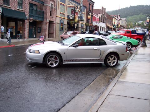 1972 Ford Ranchero - A silver coupe with red racing stripes is parked on a wet street in front of shops.