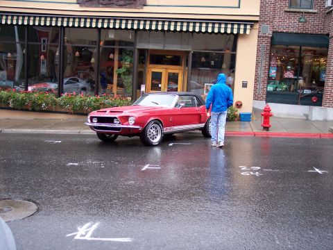 1972 Ford Ranchero - A red convertible Mustang is parked on a wet street in front of a building with awnings.