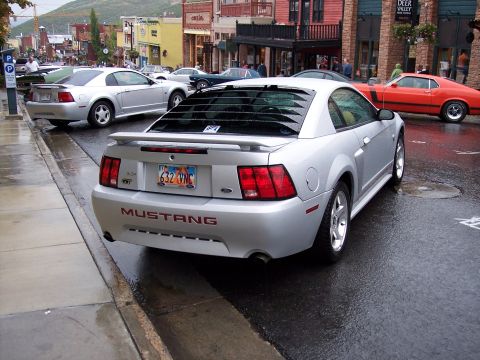 1972 Ford Ranchero - A silver Ford Mustang is parked on a wet street in front of shops.