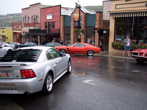 1972 Ford Ranchero - A silver Ford Mustang is parked on a wet street in front of shops, with a red Mustang visible in the background.