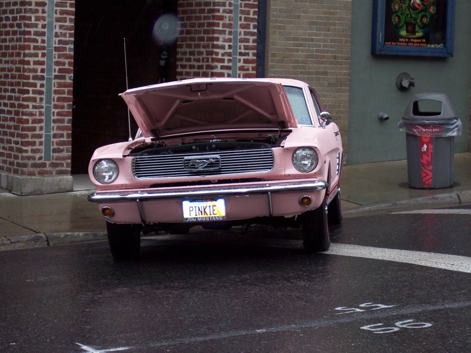Pink classic Ford Mustang, front view, hood open, 'PINKIE' plate, parked on wet street