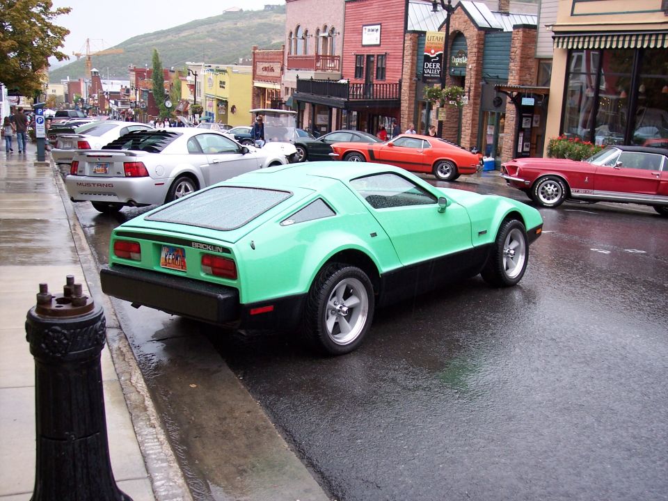 Mint green Bricklin SV-1 coupe parked on a wet street at a car show with vintage Mustangs behind.