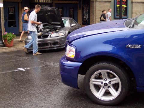 1972 Ford Ranchero - A blue Ford F-150 Lightning is parked on a wet street next to a gray car with its hood open.