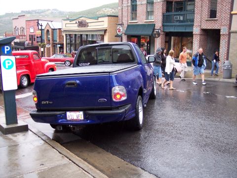 1972 Ford Ranchero - A blue pickup truck is parked on a wet street in front of shops.