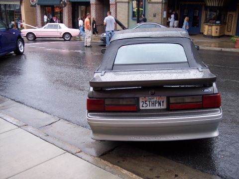 1972 Ford Ranchero - A dark gray Ford Mustang GT convertible is parked on a wet street next to a sidewalk. Another classic car is visible across the street.