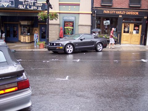 1972 Ford Ranchero - A dark gray Ford Mustang is parked on a wet street in front of a Harley-Davidson store.