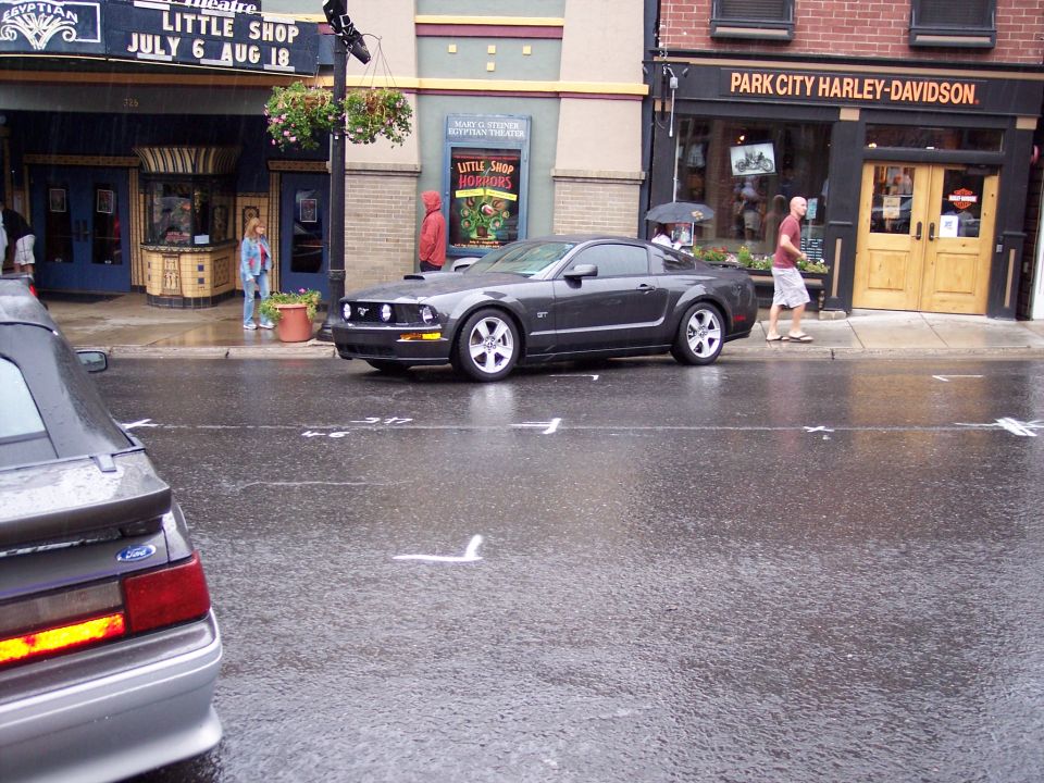 Dark gray Mustang GT coupe parked on wet street by Harley-Davidson store