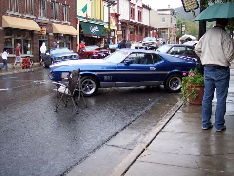 1972 Ford Ranchero - A blue 1971 Ford Mustang Mach 1 is parked on a wet street in a town, with people and other cars around.