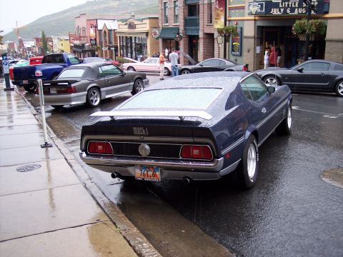 1972 Ford Ranchero - A dark blue 1971 Ford Mustang Mach 1 is parked on a wet street in front of shops.