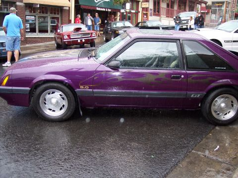 1972 Ford Ranchero - A purple Ford Mustang GT with flame decals is parked on a wet street, with other cars and buildings in the background.