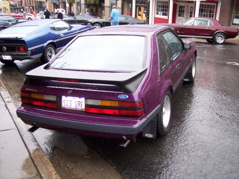 1972 Ford Ranchero - A purple 1987 Ford Mustang hatchback is parked on a wet street, with other vehicles visible in the background.