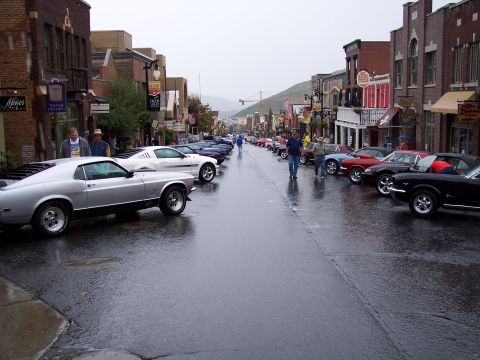 1972 Ford Ranchero - Classic cars line a wet street in a town, with buildings on either side.