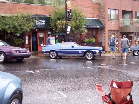 1972 Ford Ranchero - A blue muscle car with a spoiler is parked on a wet street in front of a restaurant.