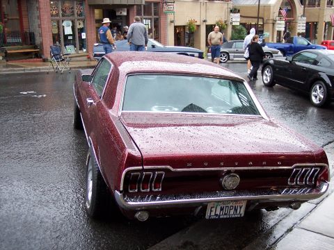 1972 Ford Ranchero - Maroon 1967 Ford Mustang coupe parked on a wet street, with other vehicles and buildings in the background.