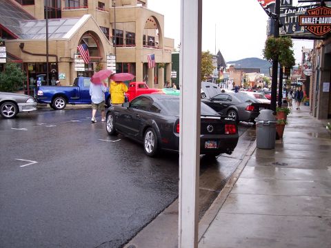 1972 Ford Ranchero - Several cars are parked on a wet street in front of shops, with people holding umbrellas.