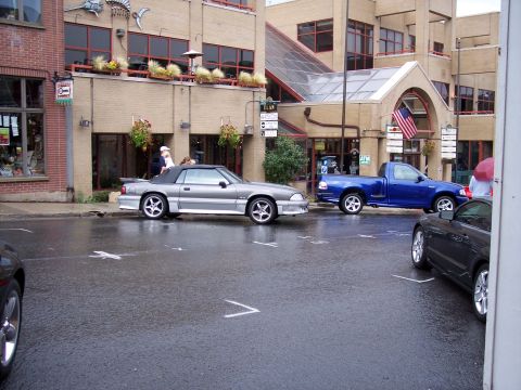 1972 Ford Ranchero - Two vehicles are parked on a wet street in front of shops on a cloudy day.