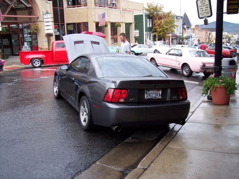 1972 Ford Ranchero - A dark gray coupe is parked on a wet street, with a red pickup truck and a pink classic car visible in the background.
