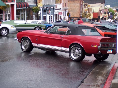 1972 Ford Ranchero - A red convertible muscle car is parked on a wet street, with other vehicles and buildings in the background.