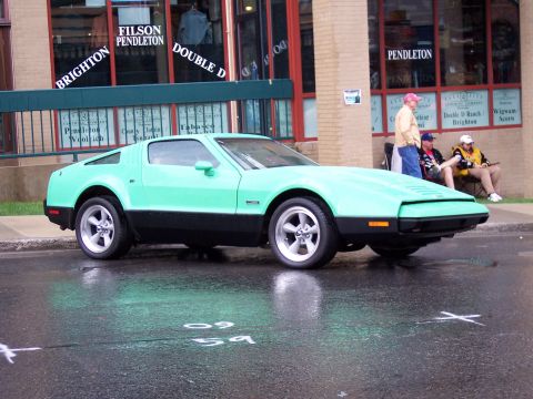 1972 Ford Ranchero - A two-tone sports car is parked on a wet street in front of a store with several people sitting nearby.