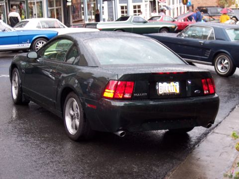 1972 Ford Ranchero - A dark green Ford Mustang Bullitt is seen from the rear on a wet street, with other vehicles in the background.