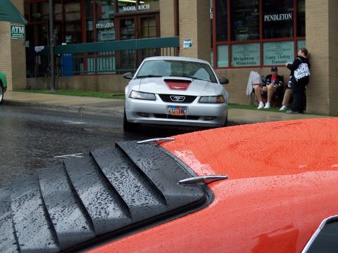 1972 Ford Ranchero - A silver Ford Mustang with a red stripe on the hood is parked on a wet street, partially obscuring an orange vehicle in the foreground.