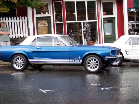 1972 Ford Ranchero - A blue 1968 Ford Mustang convertible is parked on a wet street in front of a building with shops.