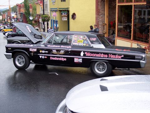 1972 Ford Ranchero - A black 1964 car, possibly a Falcon, is parked on a wet street, adorned with numerous racing decals and the prominent text "Moonshine Haule…