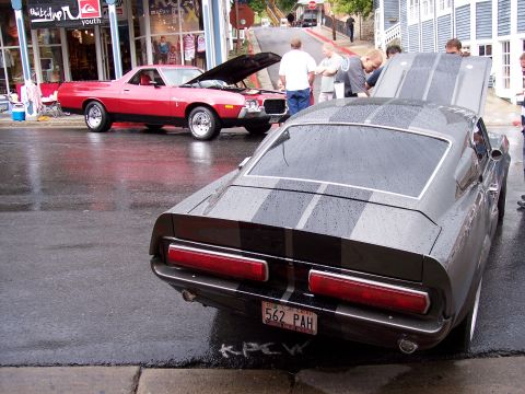 1972 Ford Ranchero - A gray 1967 Ford Mustang Fastback with racing stripes is parked on a wet street, with a red Ford Ranchero in the background.