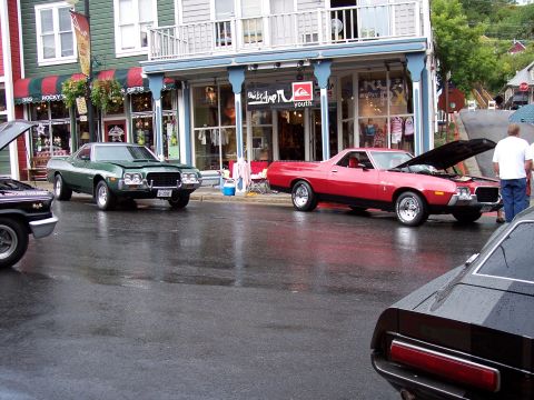 1972 Ford Ranchero - Two Ford Rancheros parked on a wet street in front of shops, one with its hood open.