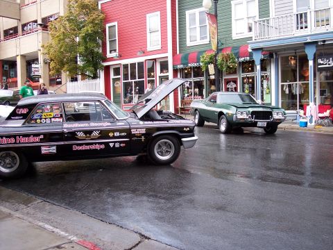 1972 Ford Ranchero - A green 1972 Ford Ranchero is parked on a wet street next to a black car with racing decals and an open hood.