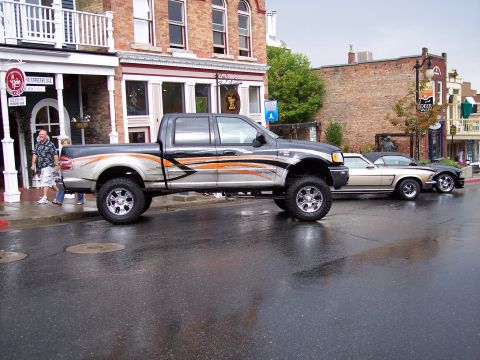 1972 Ford Ranchero - A lifted pickup truck with custom graphics is parked on a wet street in front of buildings, with classic cars visible behind it.