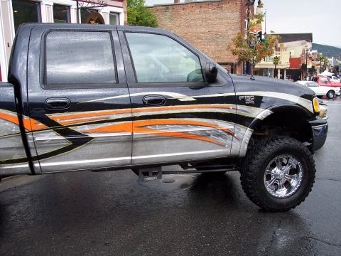 1972 Ford Ranchero - A lifted Ford F-150 pickup truck with custom graphics is parked on a wet street in front of buildings.