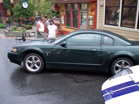 1972 Ford Ranchero - A dark green coupe with a black roof sits on a wet street in front of a building with colorful awnings.
