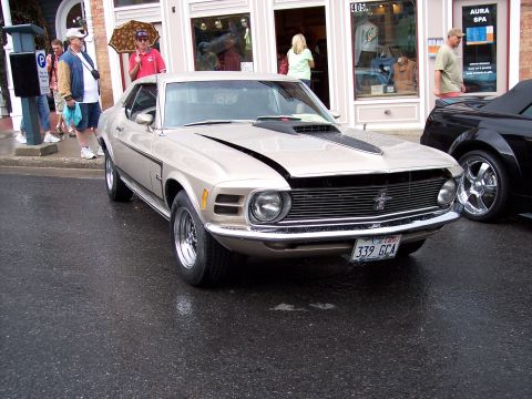 1972 Ford Ranchero - A tan 1970 Ford Mustang is parked on a wet street in front of shops, with another car visible to its right.
