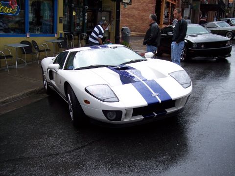 1972 Ford Ranchero - A modern Ford GT with racing stripes sits on a wet street in front of a storefront.