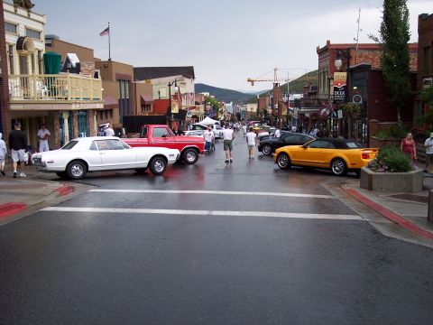 1972 Ford Ranchero - A white coupe and a red pickup truck are parked on a wet street in a town square, with other vehicles and people visible.